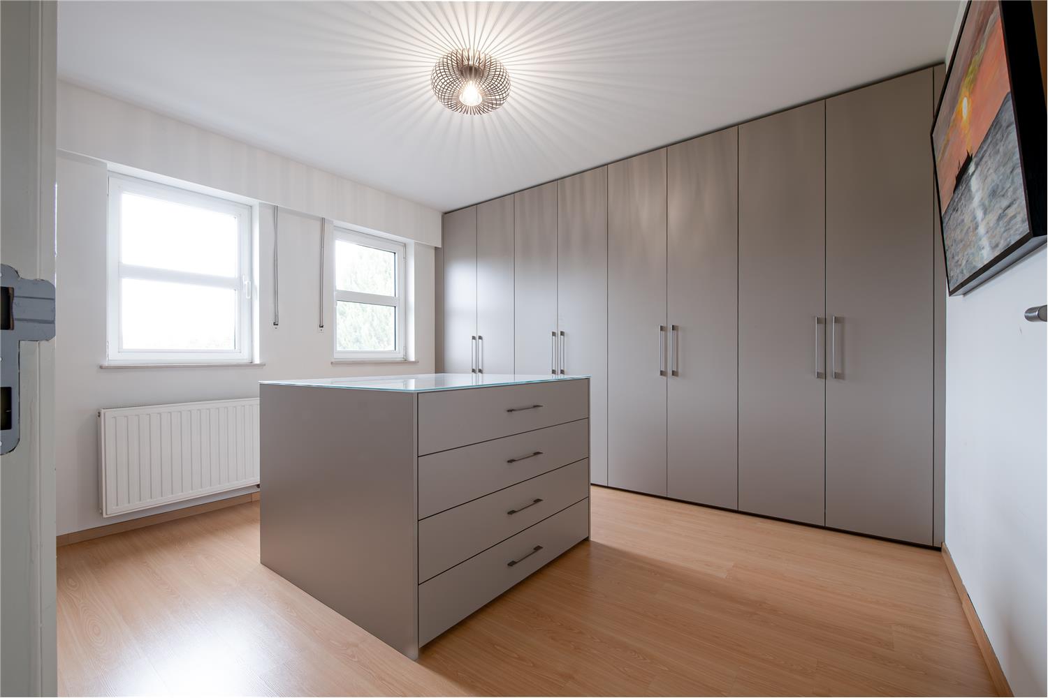 Overzicht van de dressing in Essen met kamerhoge taupekleurige maatkasten en een centraal ladeblok.
