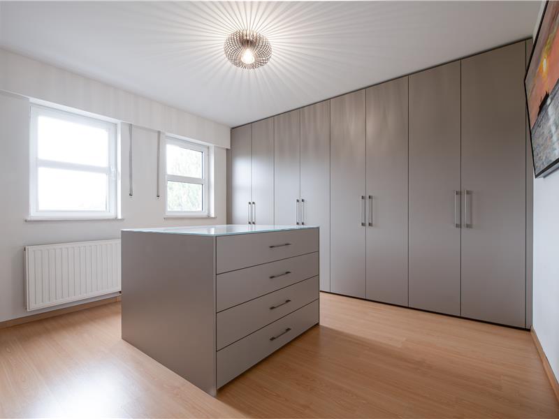 Overzicht van de dressing in Essen met kamerhoge taupekleurige maatkasten en een centraal ladeblok.